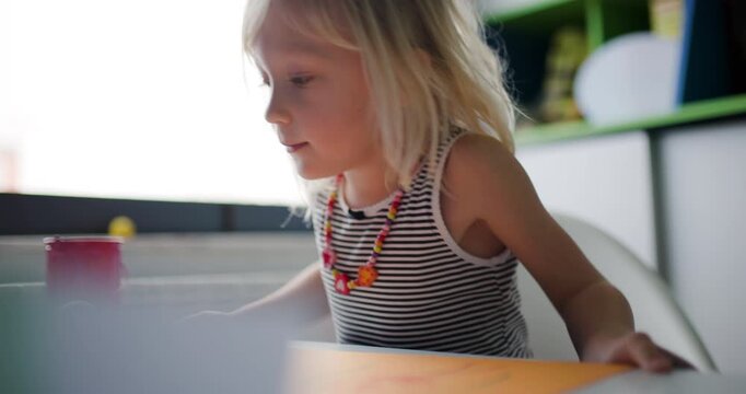 Happy Little Girl Drawing with Felt-Tip Pens on Orange Paper at Her Desk by the Window, Smiling and Enjoying Creative Activity