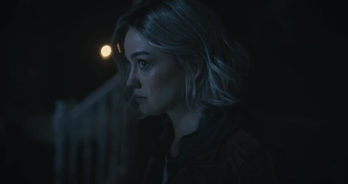 Cautious looking young woman staring around dark home as shadowy figure walks behind her then staring down at her notebook with flashlight - closeup tilt shot