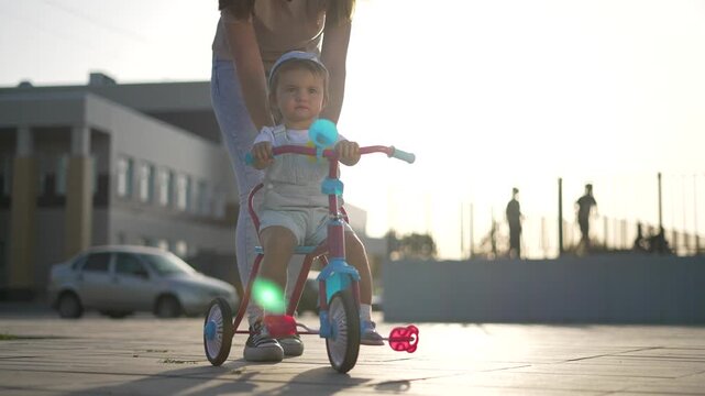Mother pushing toddler on tricycle. Young mother helps toddler riding in city plaza. Family enjoys outdoor play at sunset. Pushing tricycle brings family joy. Bright sunset warms paved plaza.