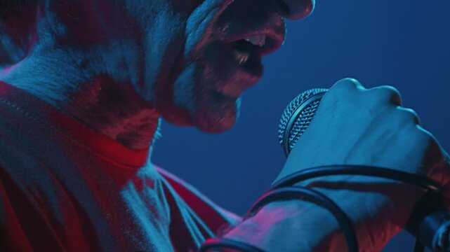 Close up of male hand holding microphone. Black cable wrapped around arm. Energetic live music performance with dramatic pink, blue, purple stage lighting. Raw, intense atmosphere of hard rock show