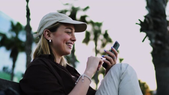 Joyful female traveler wearing baseball cap laughing during smartphone chat outdoors. Cheerful woman content creator watching multimedia video online connection workspace background
