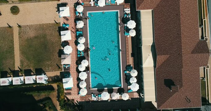 Aerial top down view of a rectangular blue swimming pool surrounded by white umbrellas and sun loungers at a resort