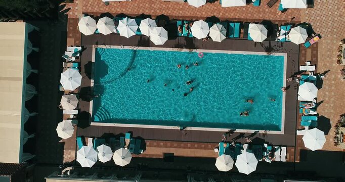 Aerial top down view of a rectangular blue swimming pool surrounded by white umbrellas and sun loungers at a resort