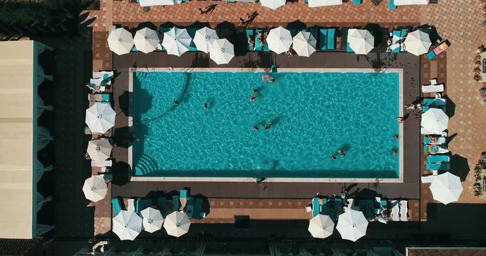 Aerial top down view of a rectangular blue swimming pool surrounded by white umbrellas and sun loungers at a resort