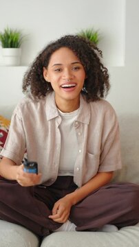 Vertical screen, Young African American woman with curly hair sitting cross-legged on sofa, using a TV remote. Calm leisure lifestyle in a bright modern living room with plants.