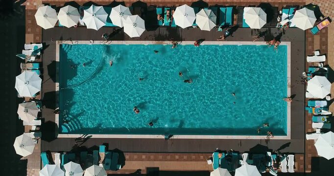 Aerial top down view of a rectangular blue swimming pool surrounded by white umbrellas and sun loungers at a resort