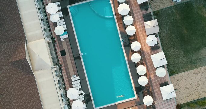 Aerial top down view of a rectangular blue swimming pool surrounded by white umbrellas and sun loungers at a resort