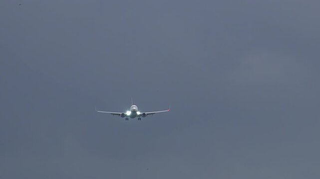 Narrow body jet on final approach with bright landing lights glowing, gear extended, front view centered in frame against dark dramatic overcast sky