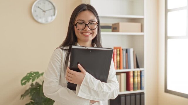 Woman holding black binder to chest in building with bookshelves, clock, plant and window visible, smiling and wearing glasses; confidence study focus.