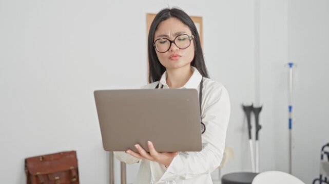 Woman doctor with stethoscope holds laptop and furrows brow while reading patient notes in clinic; concern focus.