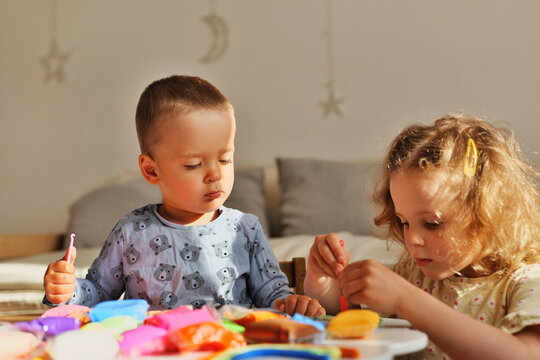 Two young children engage in creative play with colorful clay on a table. Sunlight fills the room, creating a happy atmosphere for their activity