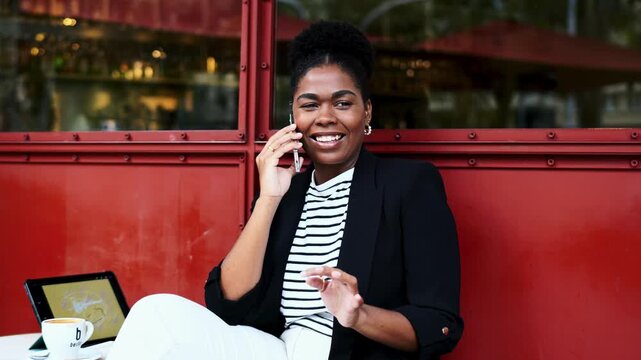 Happy multiracial young businesswoman cellular conversation modern cafe terrace freelance work. African American female entrepreneur laughing negotiating online business transaction
