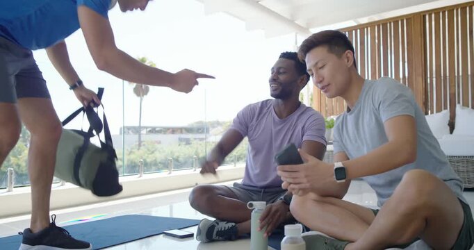 Diverse male friends viewing phone, planning workout on porch while newcomer arriving with duffel