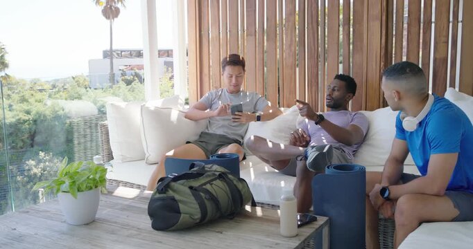 Diverse male friends on patio sofa, left man scrolling phone, middle man pointing, planning workout