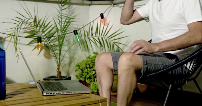 Balcony Oasis: A man relaxes on a comfortable chair on a balcony, laptop open beside him, surrounded by lush plants and festive string lights, creating a peaceful work or leisure space.
