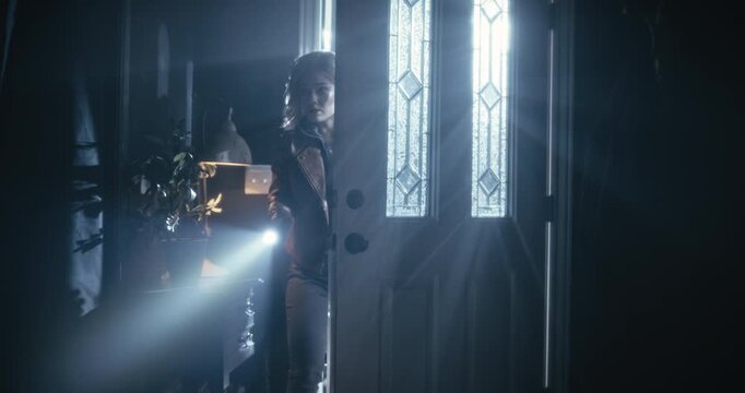 Silhouette of young woman with flashlight entering dark home with cautious look - dolly in