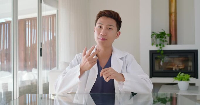 Asian man in white coat addressing camera, gesturing to chest, explaining health at glass table