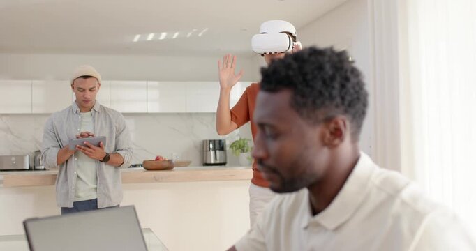 Diverse male team working in home kitchen, foreground man typing on laptop while monitoring VR demo
