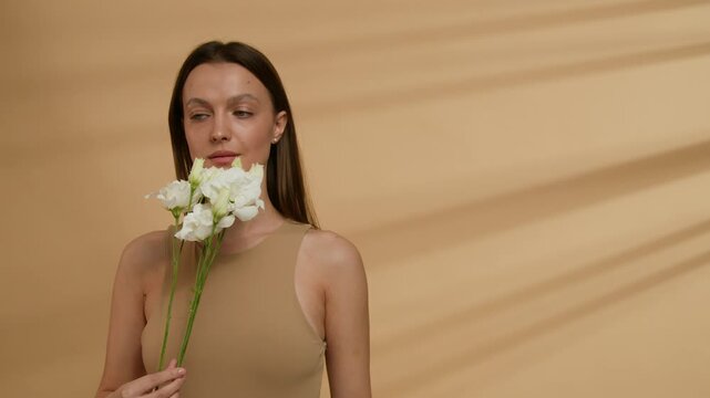 Waist-up view of Caucasian young woman dressed in beige tank top holding small bouquet of eustoma flowers in studio