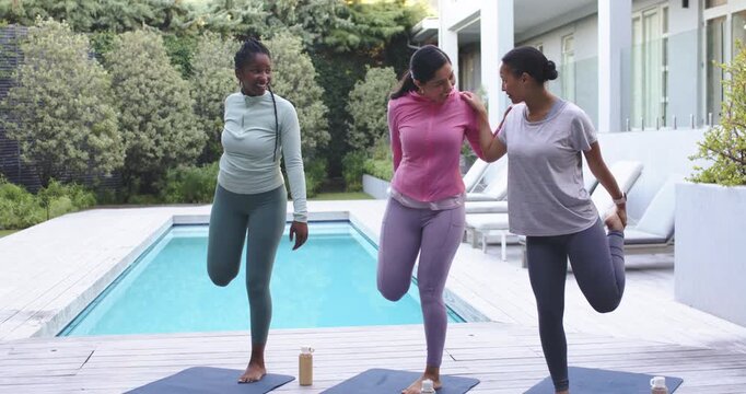 Diverse women in leggings holding ankles on yoga mats by pool, center steadying right for balance