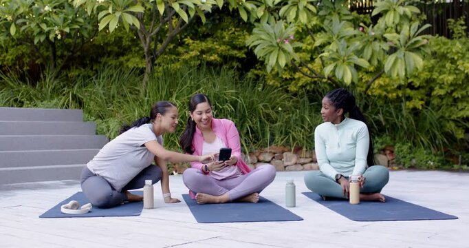Diverse women in activewear sitting on mats at patio, center tilting smartphone, sharing, laughing