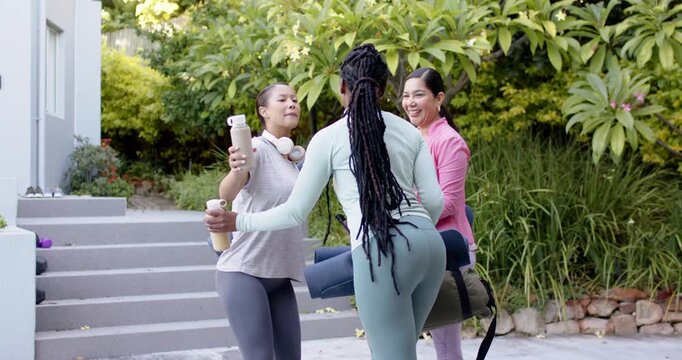 Diverse female friends meeting at front steps in gymwear with mats and bottles hugging for workout