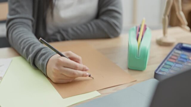 Woman wearing glasses drawing with pencil, hand visible sketching on paper beside watercolor palette, pencil case and wooden mannequin in studio; concentration creativity.