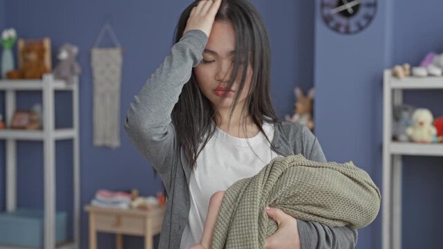 Young woman cradling swaddled baby with hand on forehead, eyes closed, holding blanket in a room inside a building; maternal exhaustion.
