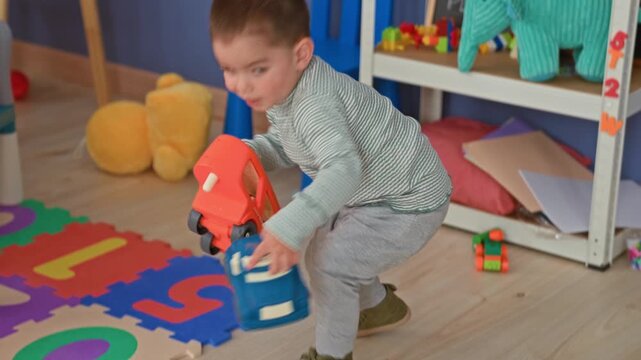 Boy crouching and holding toy cars, pushing a blue car across foam alphabet mat beside a shelf and stuffed elephant in a playroom; happy curiosity.