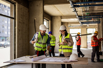Male and female architects reviewing renovation blueprints with builders working in the background. © Mediteraneo