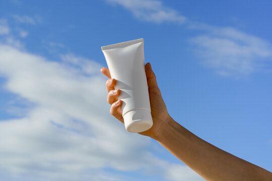 A hand holds a white mockup tube against a bright blue sky. Blank unbranded container for cosmetic product mockups like sunscreen or lotion. Skincare, sun protection, and summer vacation concepts