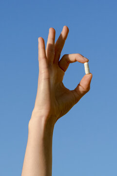 Hand holding a white mockup pill in an OK gesture against a blue sky. Concept for health, wellness, and successful treatment. Minimalist composition with bright light and ample blue copy space