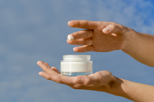 Hands holding a glass jar of white mockup moisturizer. A dab of cream is on the fingertrypsin, ready for application. Natural daylight a blue sky create a fresh, clean mood for skincare beauty concept