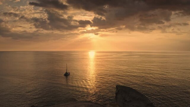 Sunset paints sky and ocean in warm golden hues, with sun rays reflecting on the tranquil water surrounding a serene catamaran, viewed from a rocky Phuket island coastline. Aerial view drone flight