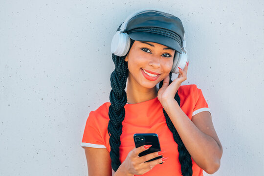 happy young woman listens to music on white headphones while holding a phone. She wears a cap and long braids