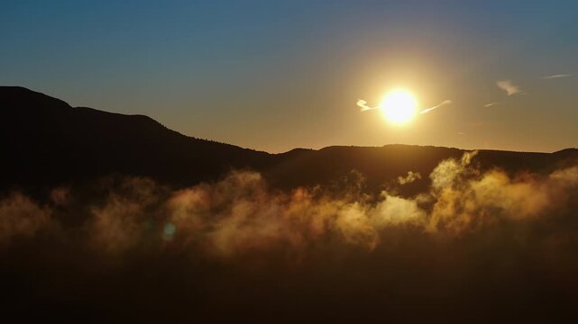 Wide aerial view of mountain ridge at sunrise with a broad golden fog layer drifting low across terrain, bright sun rising under clear blue sky right of frame