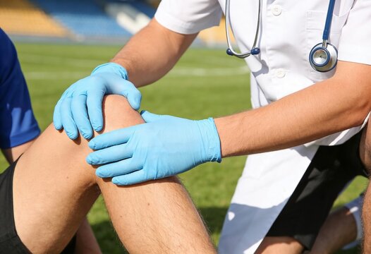 A sports physician examines a soccer player's injured knee while on the grass of a sports field.