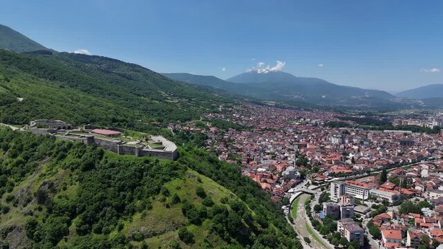 Aerial drone footage of Prizren city and its historic fortress captured on a clear sunny day, highlighting the beautiful blend of traditional architecture, urban life, and the famous hilltop castle ov