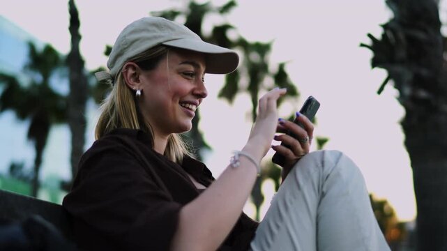 Smiling female digital nomad wearing cap checking mobile phone screen notification outdoors. Happy woman traveler browsing social media network connection remote office palm trees view
