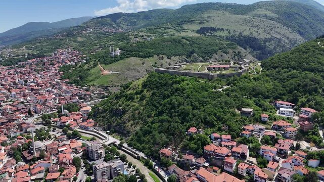 Aerial drone footage of Prizren city and its historic fortress captured on a clear sunny day, highlighting the beautiful blend of traditional architecture, urban life, and the famous hilltop castle ov