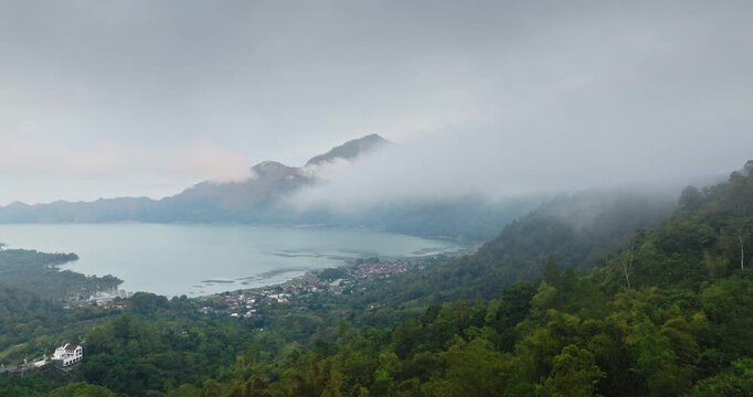 Mount Batur caldera, Lake Batur and Kintamani village below, enveloped by lush tropical forests and dramatic clouds creating mystical atmosphere over Bali volcanic landscape. Aerial view drone flight