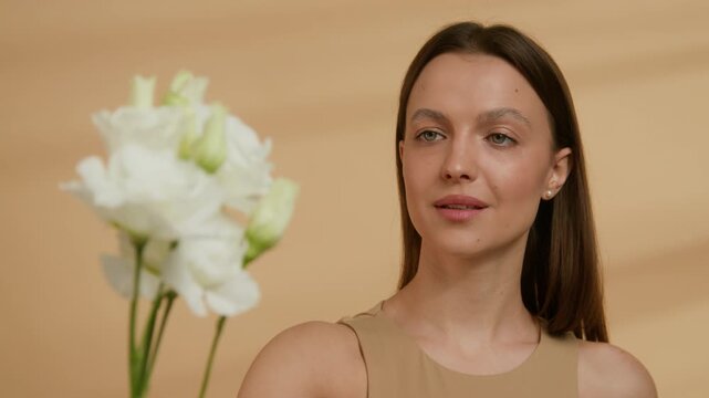 Rack focus shot of Caucasian girl looking at bouquet of white eustoma flowers with admiration against beige studio background