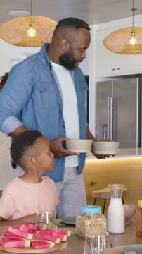 Vertical video: Mom placing watermelon at counter dad in denim holding bowls serving breakfast