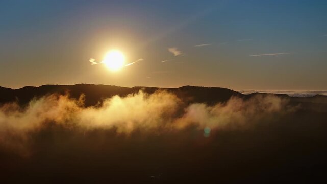 Dense golden fog bank fills the lower frame beneath a dark mountain silhouette, sun above the ridge with blue sky overhead and warm light saturating the mist