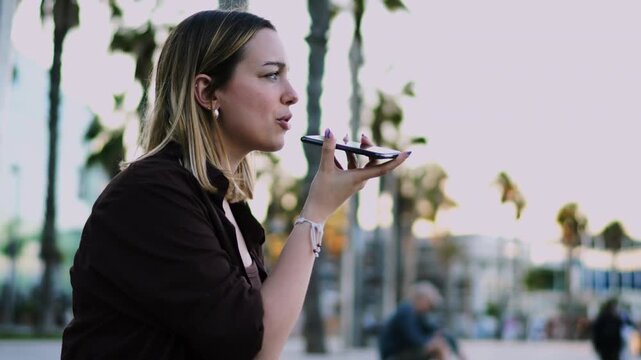 Laughing female student recording voice message holding smartphone speaker outdoor profile view. Happy woman freelancer talking mobile phone conversation online communication lifestyle
