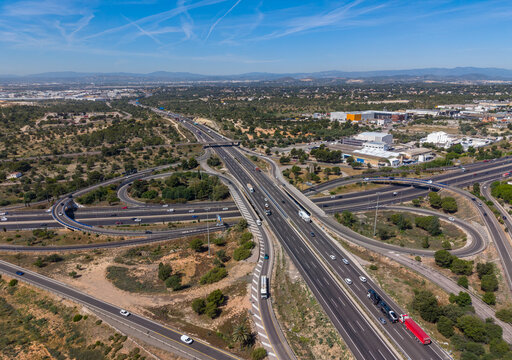 Aerial view of a multilane cloverleaf highway interchange near Valencia, Spain, with heavy mixed traffic including a red articulated lorry, set against a Mediterranean landscape
