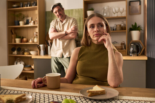 Adult woman sitting thoughtfully at table while man standing behind with crossed arms, showing relationship tension. Useful for conflict counseling or family stress themes