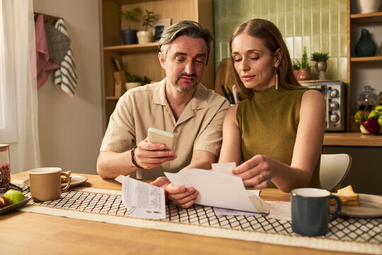 Couple reviewing household bills, checking smartphone, managing budget at home. Useful for personal finance, banking app, debt planning, cost control