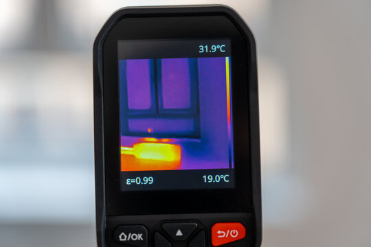 Handheld thermal camera shows room temperature data. Screen displays and with color-coded heat map. Yellow spot on floor indicates a warm area