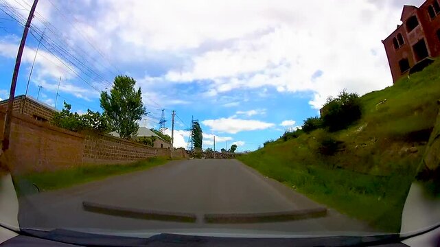Dashcam view of a car driving along an asphalt road in a rural Armenian village with stone walls and green landscape under a bright blue sky with clouds.